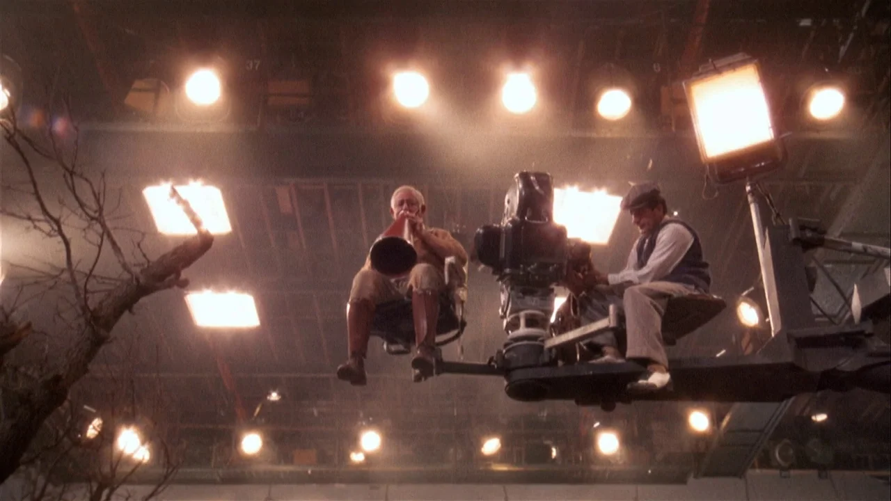 The Day of the Locust (1975) movie still: wide — A high-ceilinged film studio filled with bright overhead lights and industrial equipment. Two men…