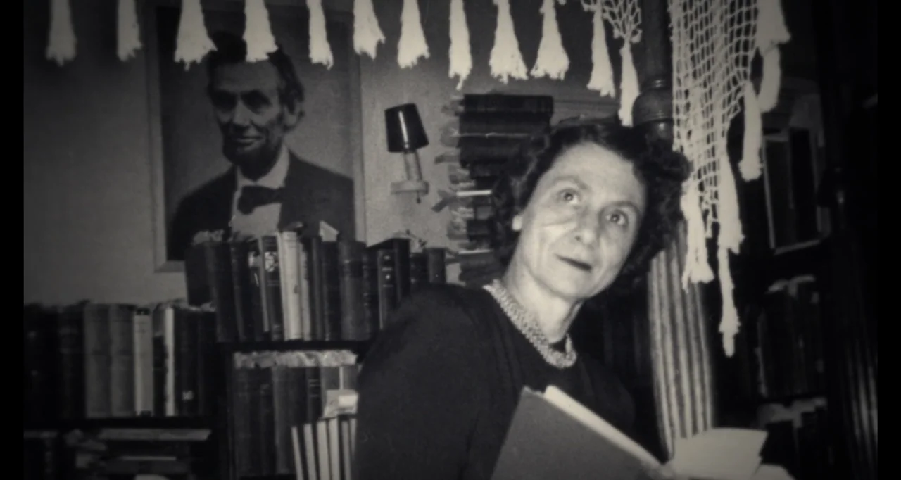 Living With Lincoln (2015) movie still: medium — A cluttered room contains bookshelves and a framed portrait of Abraham Lincoln. A middle-aged woman…