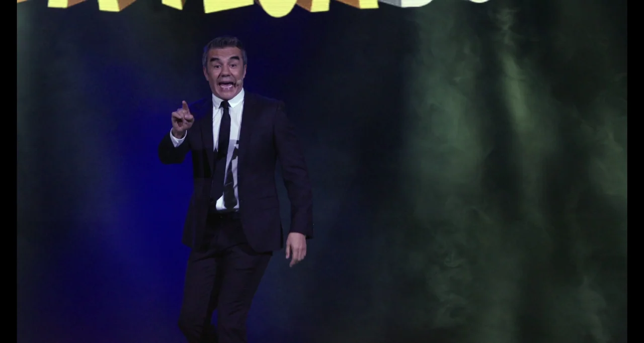 Paired Up (2019) movie still: medium — A man in a formal dark suit stands on a brightly lit stage. He points his finger while speaking to an unseen…