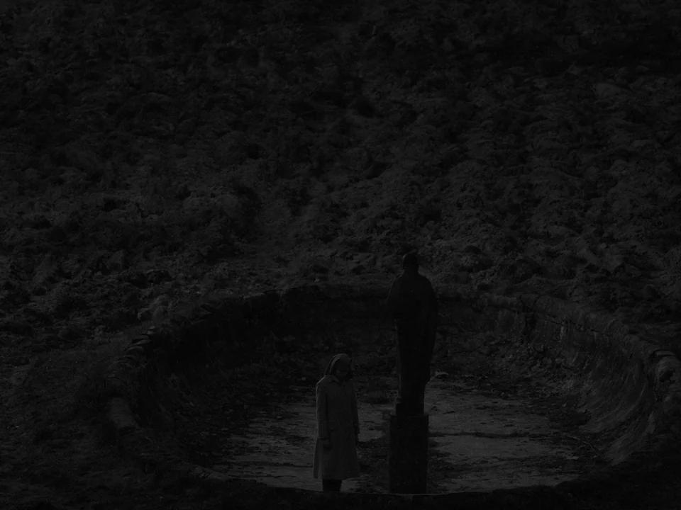 Ida (2013) movie still: wide — A dark, empty circular stone fountain sits in a desolate landscape at night. A lone nun in a long coat stands beside…