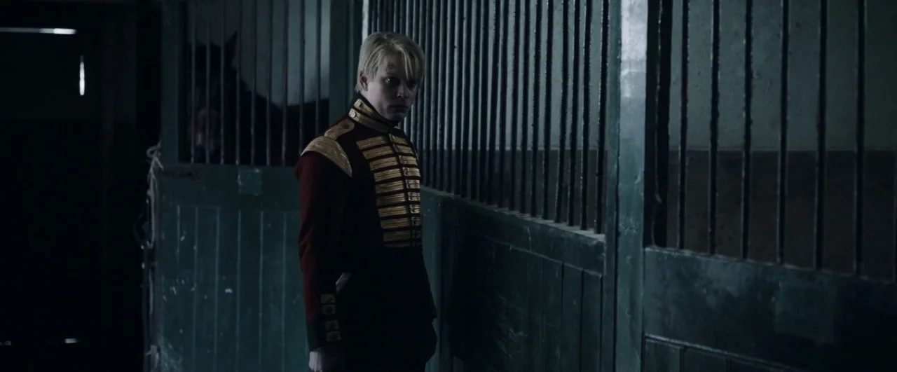 Black ’47 (2018) movie still: medium — A dark stable with green vertical bar partitions separating the interior walking area. A young man in a red…