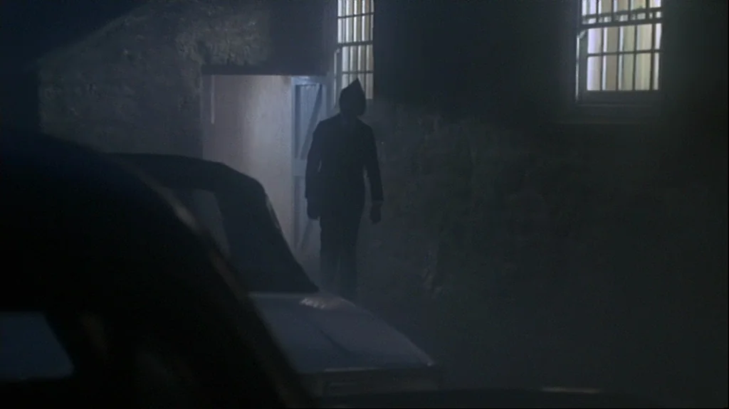 Straw Dogs (1971) movie still: fullbody — A stone building exterior at night features a glowing open doorway. A dark, silhouetted figure in a suit…