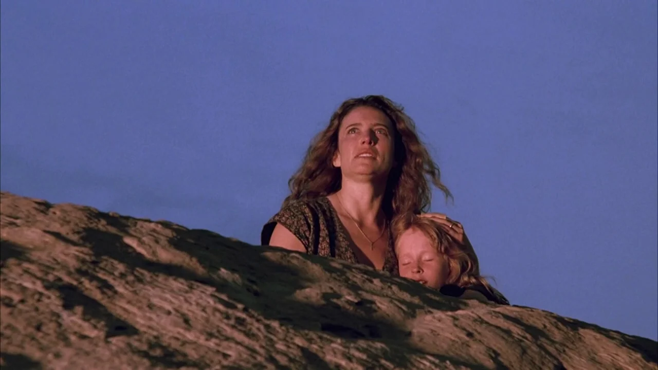 The Rapture (1991) movie still: medium — A woman and a young girl rest behind a large textured rock in a desert. The sky behind them is a deep…