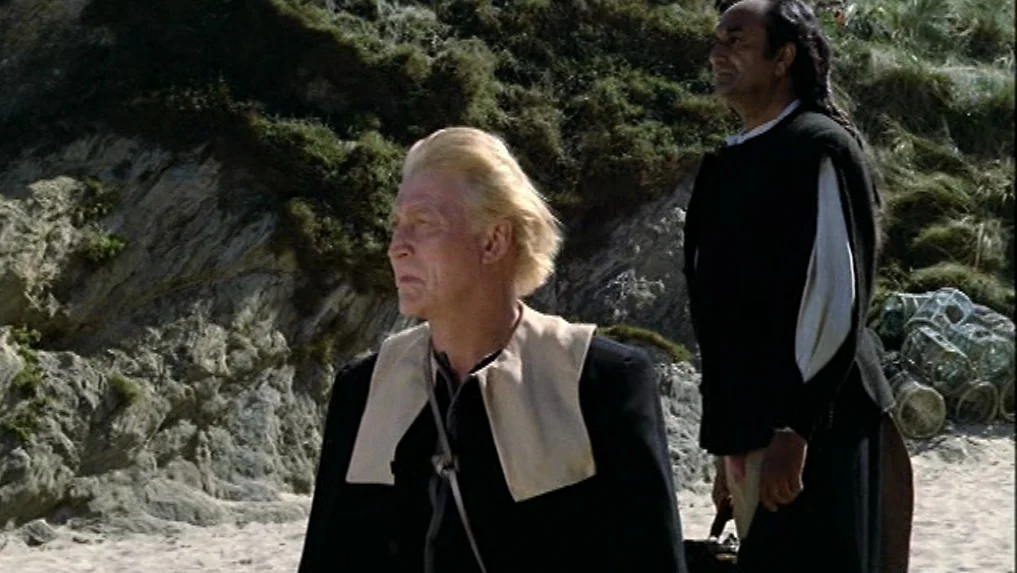 The Scarlet Letter (1973) movie still: medium — A sandy beach meets a steep, rocky cliff covered in patchy green vegetation. Two older men stand in…