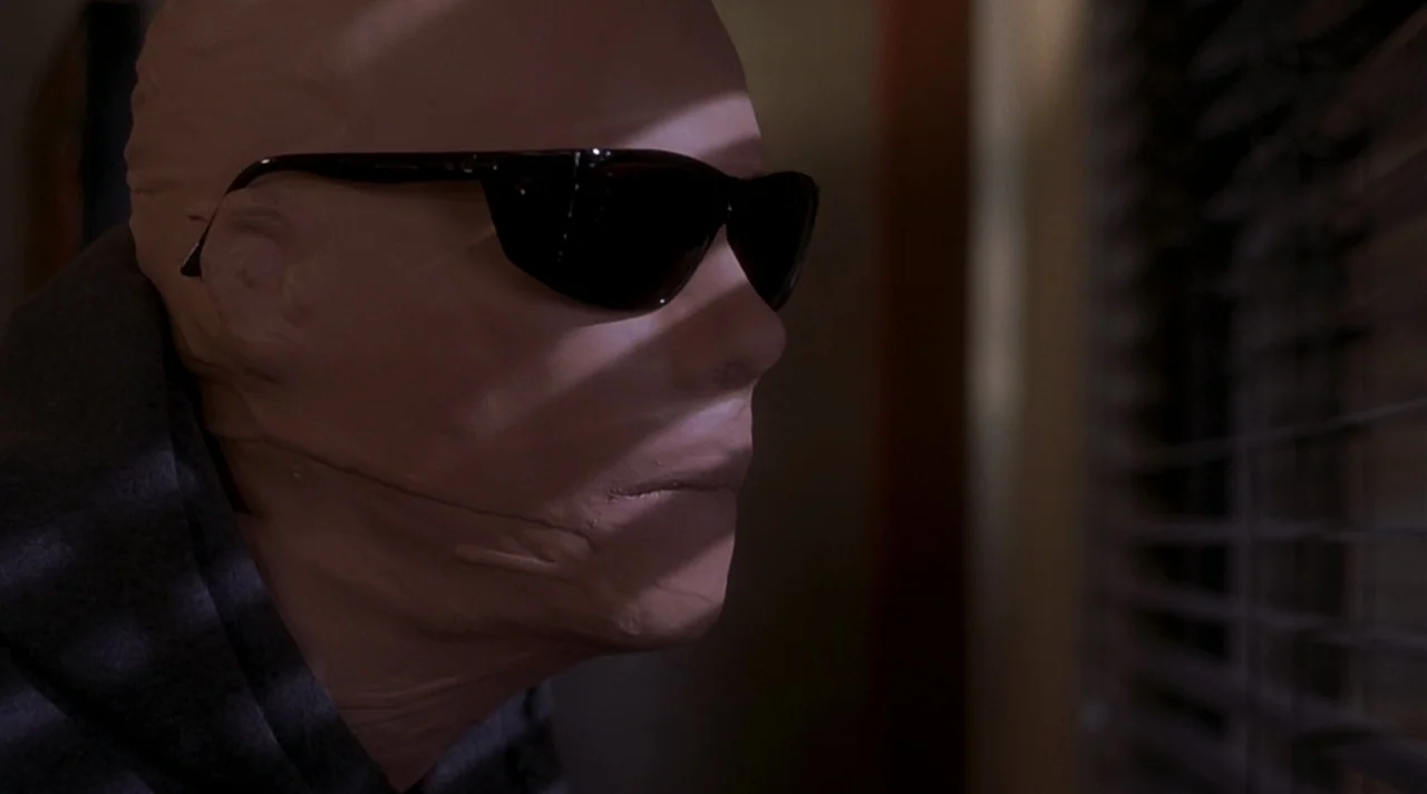 Hollow Man (2000) movie still: closeup — A figure wearing a smooth synthetic face mask peers out through closed window blinds. Hard light creates…