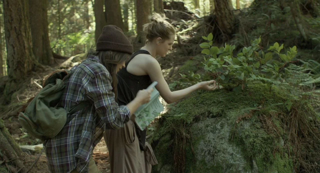En lo profundo del bosque movie still: medium — Two women stand in a dense forest with moss-covered ground and tall evergreen trees. One woman…