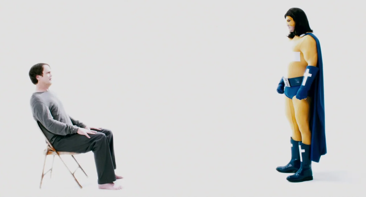 Super (2010) movie still: fullbody — A stark, brightly lit white cyclorama wall fills the entire background. A man sits on a metal folding chair…