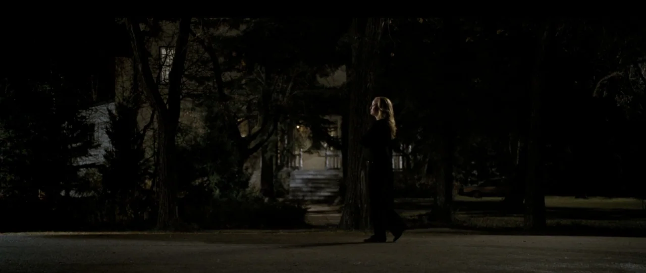 The Haunting in Connecticut (2009) movie still: fullbody — A suburban street at night with mature trees and a house in the distance. A woman stands…