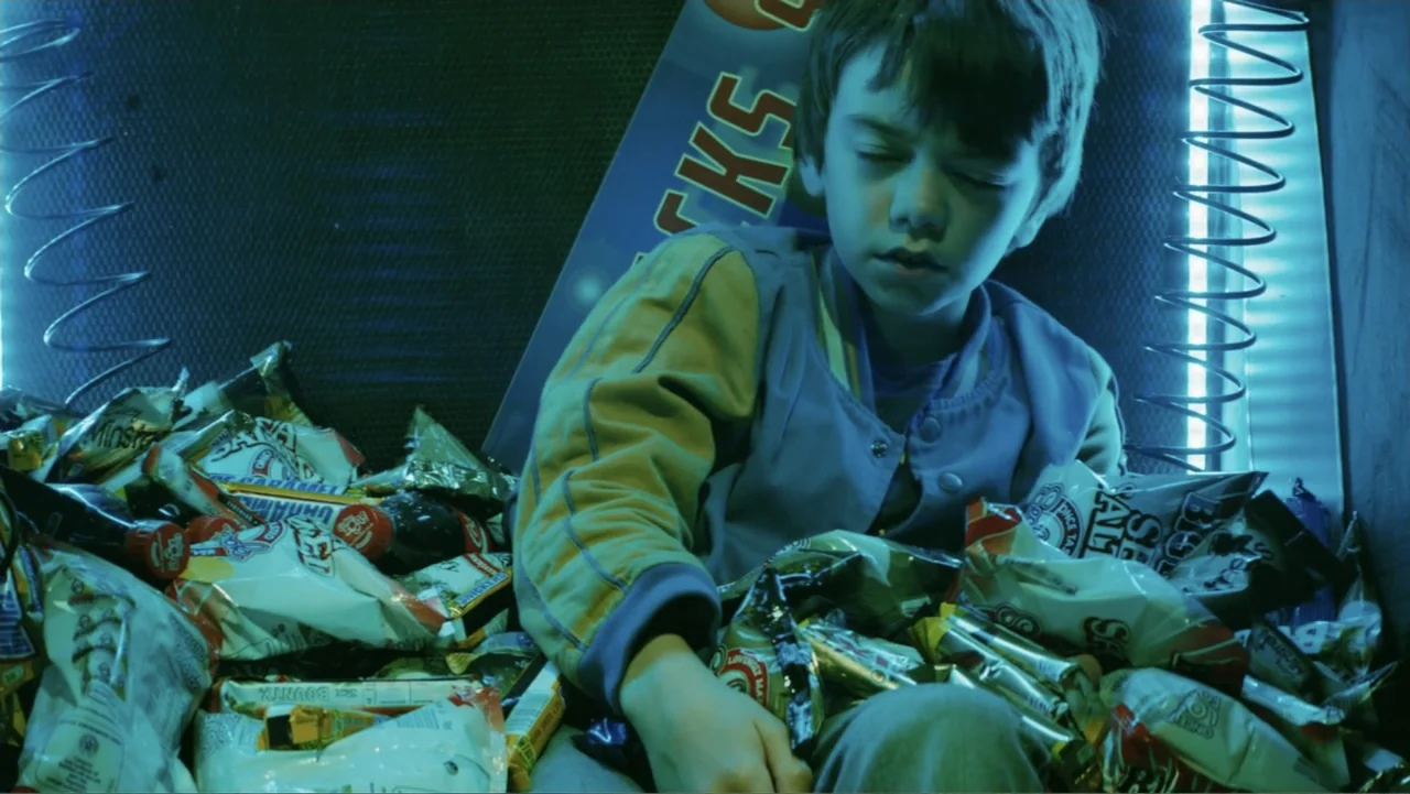 Robot Overlords (2015) movie still: medium — A young boy sits surrounded by various snack bags inside an enclosed, metallic space. The dim interior…
