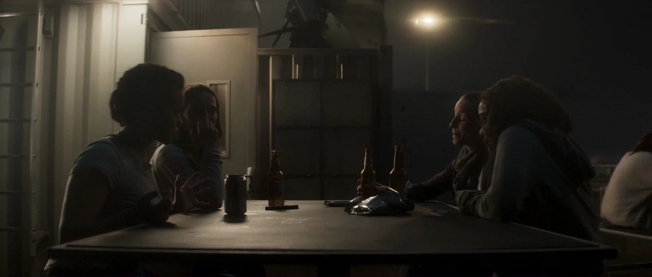 Annihilation (2018) movie still: medium — Four people sit at a table on a dark rooftop at night. They are gathered around beer bottles and a bag of…