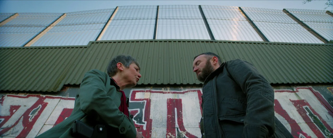 Kneecap (2024) movie still: medium — A high security fence with graffiti sits behind two people in conversation. They are framed from a low angle…
