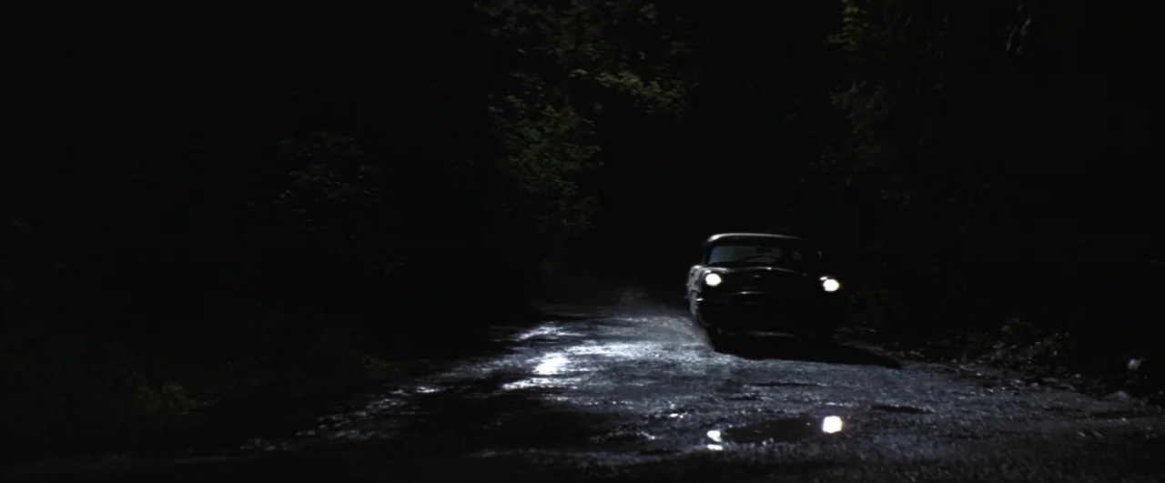 This Boy's Life (1993) movie still: wide — A dark, unpaved road winds through a densely wooded forest at night. A vintage car drives towards the…