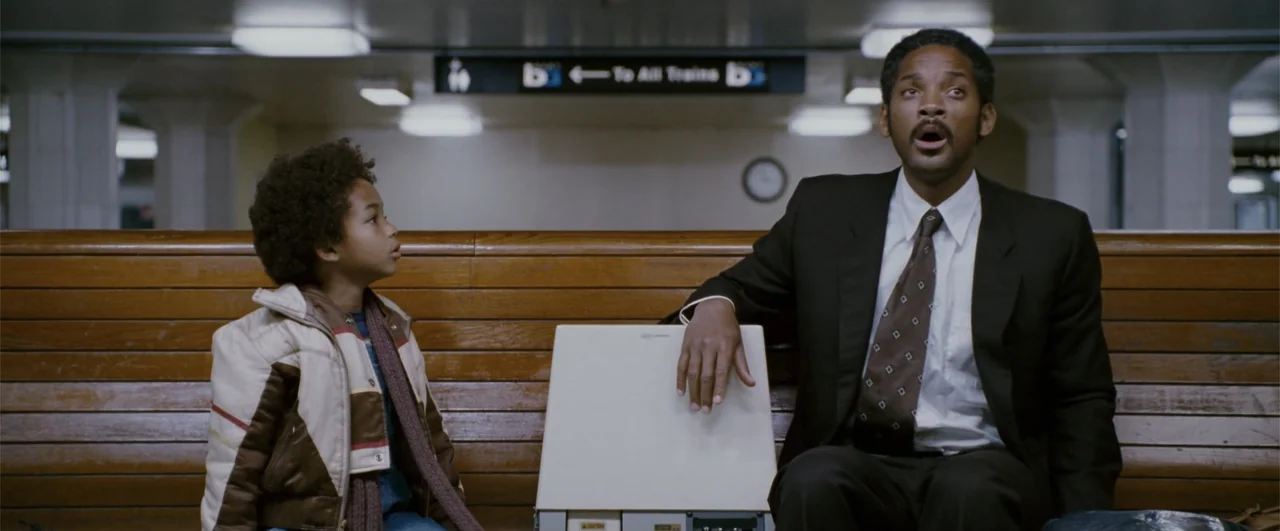 The Pursuit of Happyness (2006) movie still: medium — A tiled subway station with a brown wooden bench under ceiling lights. A man and a young boy…