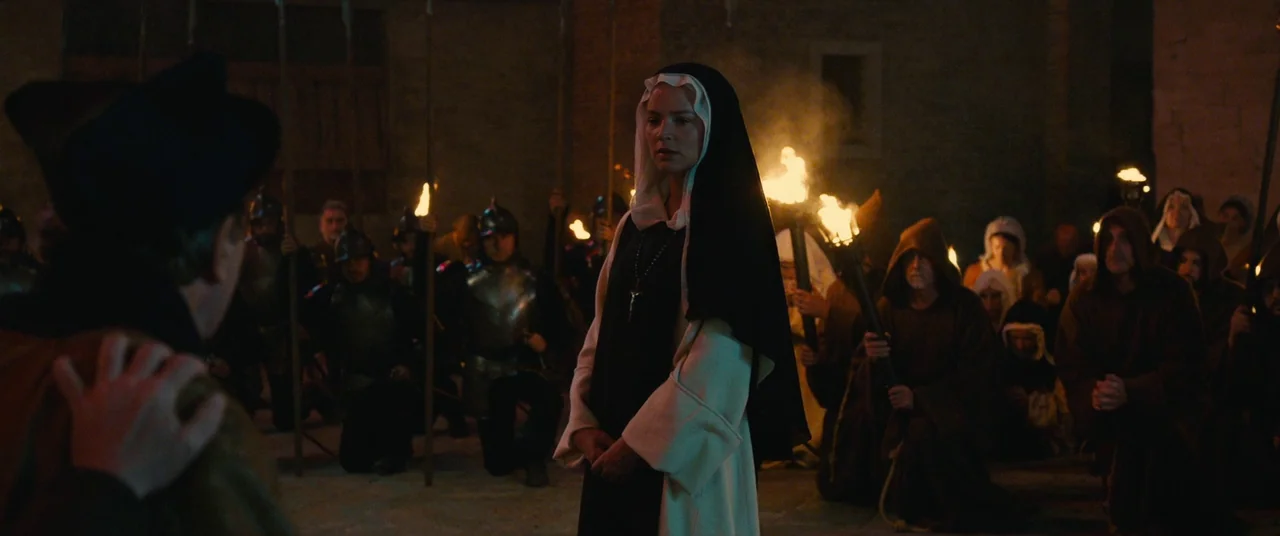 Benedetta (2021) movie still: medium — A dark night-time medieval courtyard is crowded with armed soldiers and cloaked figures. A nun stands in the…