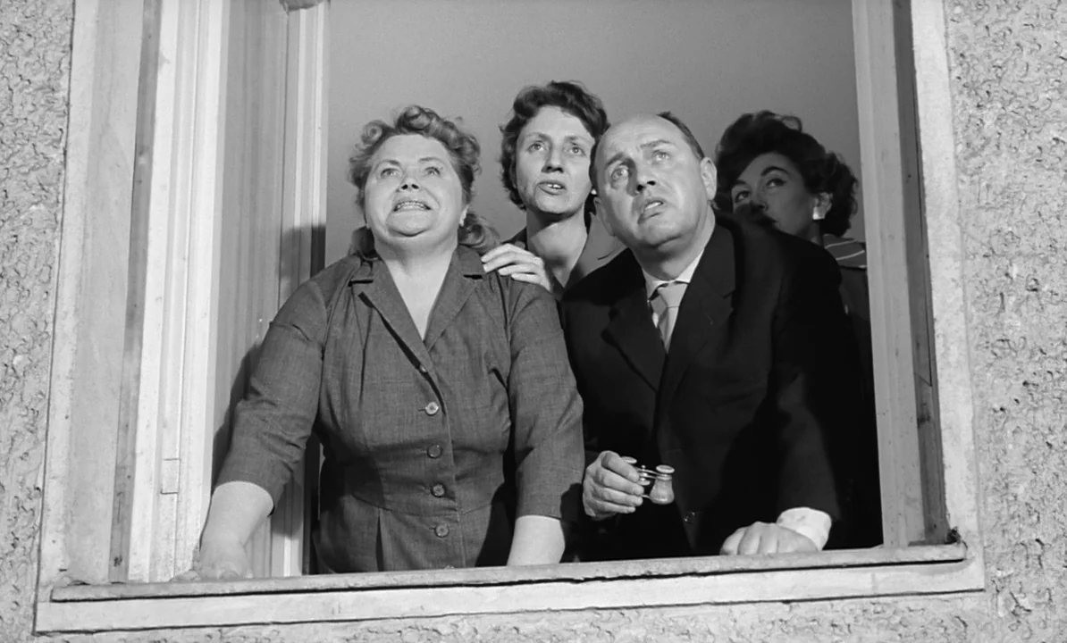 The 1,000 Eyes of Dr. Mabuse (1960) movie still: medium — A group of people watch something from a high apartment building window frame. The…