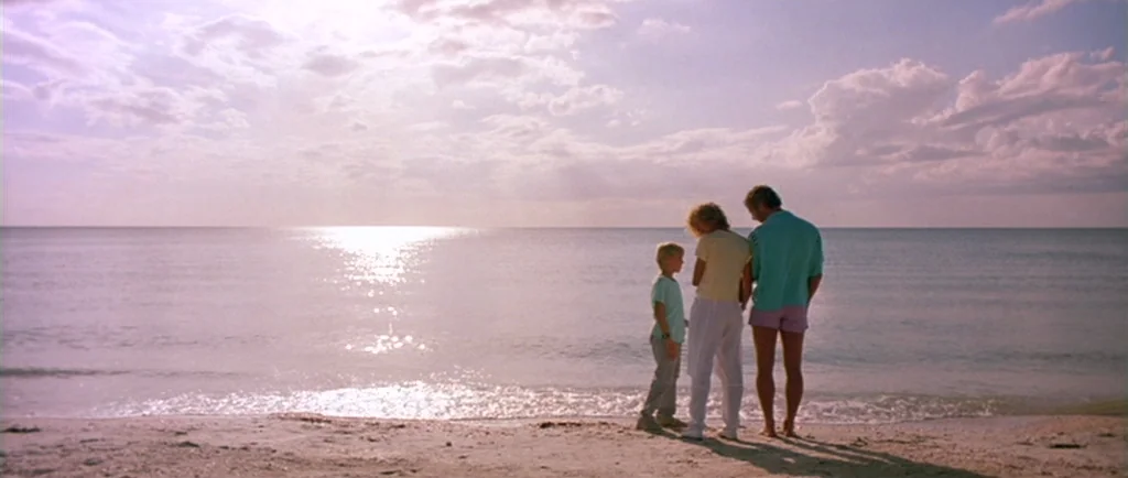 Manhunter (1986) movie still: wide — A wide beach features three people standing at the edge of the calm water. The sun hangs low in the bright,…