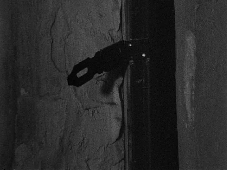 Family Nest (1979) movie still: closeup — A metal latch plate is fixed to a coarse, textured stone or concrete wall. The hardware protrudes from the…
