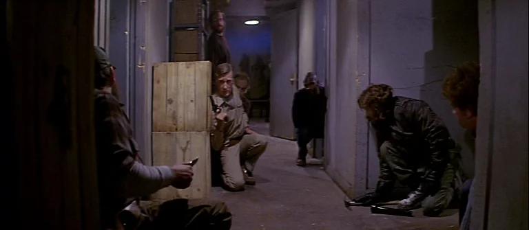 The Thing (1982) movie still: wide — A dimly lit narrow corridor features plain walls and stacked wooden crates. Several men crouch in tactical…