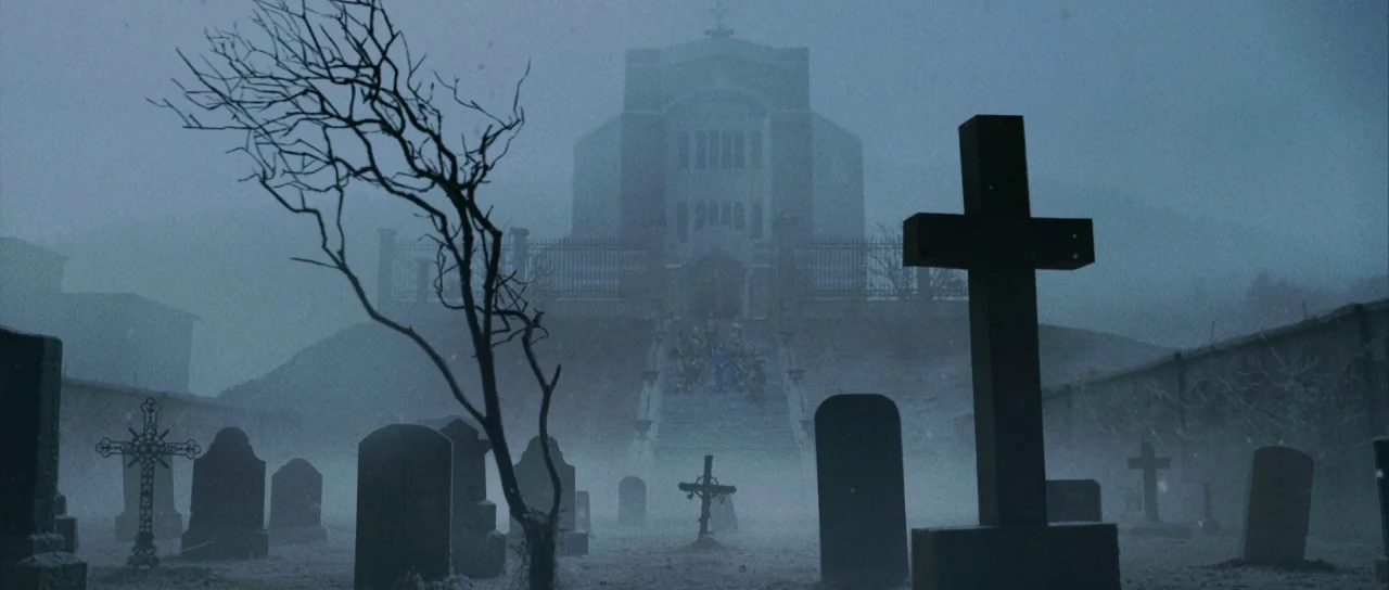 Silent Hill movie still: establishing — A dense cemetery sits under a heavy blue twilight fog with many stone markers. A large, dark building stands…