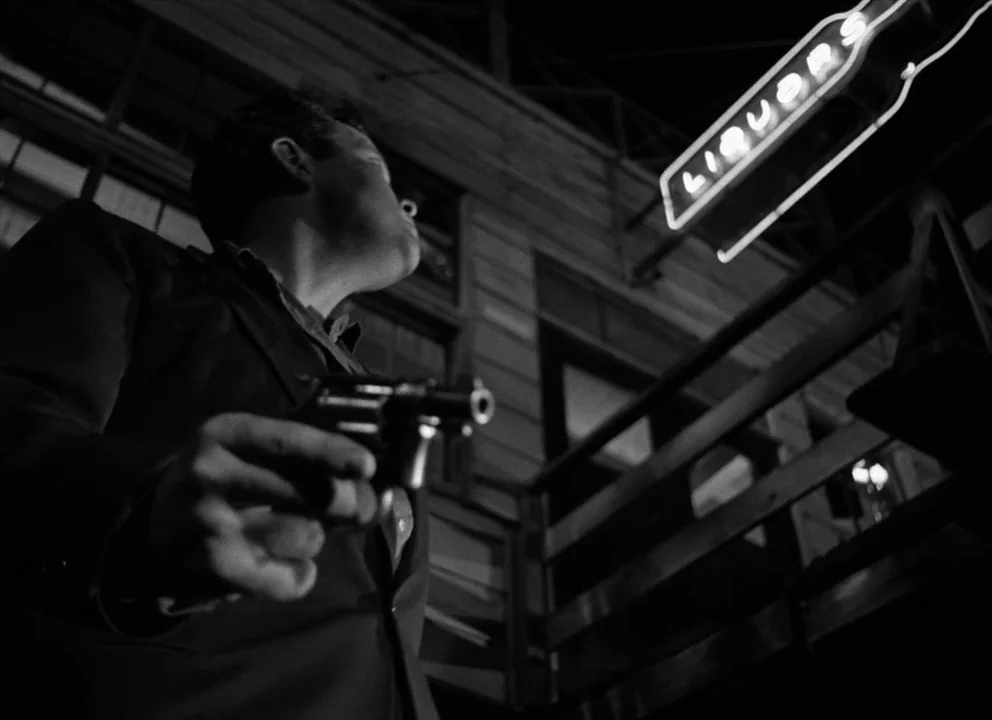 The Lady from Shanghai (1947) movie still: medium — A dark night exterior features the side of a building with a neon liquor sign. A man stands in…