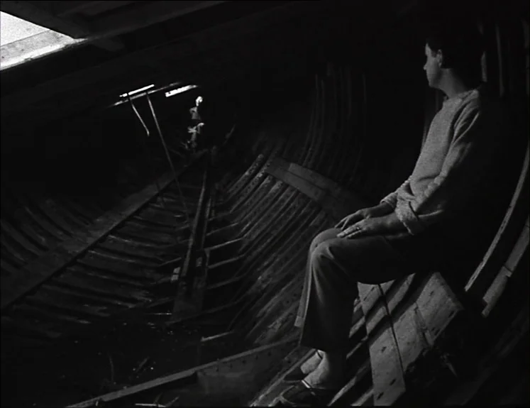 La Pointe Courte (1955) movie still: medium — The interior of a large wooden boat hull with curved structural ribs and floorboards. A man sits on a…