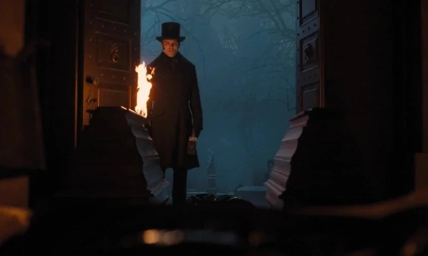 Nosferatu (2024) movie still: fullbody — A man in a top hat stands in an open doorway of a stone building. Behind him is a foggy, dark cemetery with…