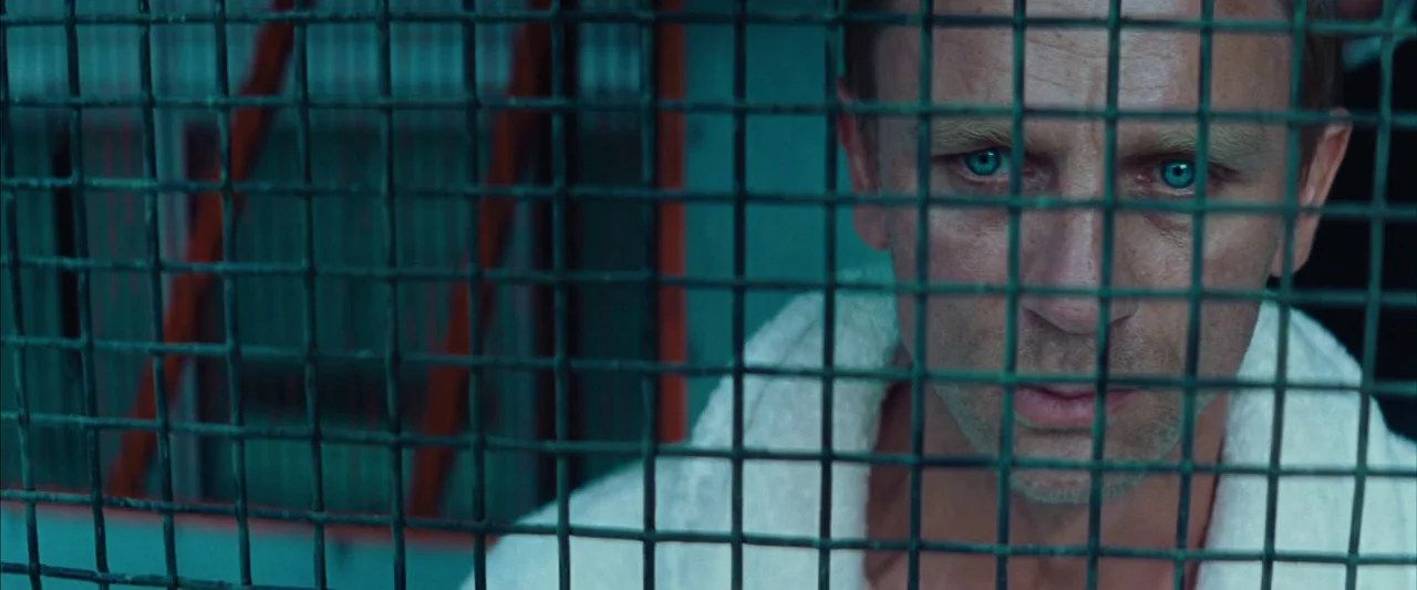 Layer Cake (2004) movie still: closeup — A man looks directly at the camera through a thick metal wire grid. The background is dark and out of focus.