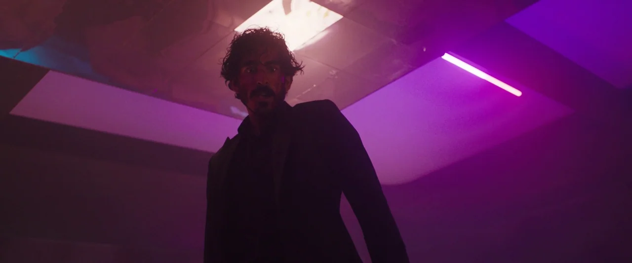 Monkey Man (2024) movie still: medium — A dimly lit indoor corridor is saturated in intense purple light from neon tubes. A man in a dark suit…