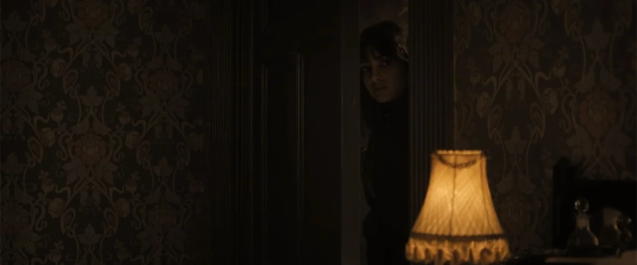 Abigail (2024) movie still: medium — A dimly lit room with vintage patterned wallpaper features a woman standing in a doorway. She is looking into…