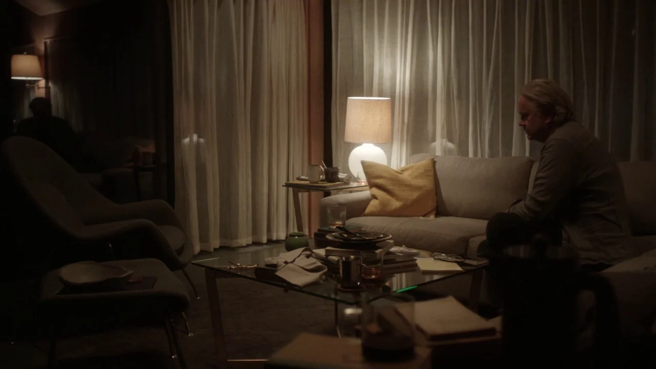 Marjorie Prime (2017) movie still: medium — A dimly lit room with sheer curtains covering large windows behind a modern sofa. A middle-aged man sits…