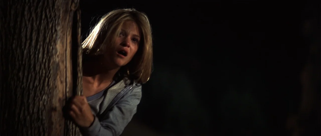 Jeepers Creepers 2 (2003) movie still: medium — A woman stands beside a large tree trunk in a dark outdoor environment at night. She peers around…