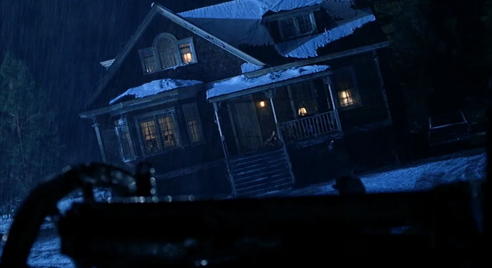Misery (1990) movie still: establishing — A wooden house with a porch stands under heavy rain at night. Snow lies on the roof while warm light glows…