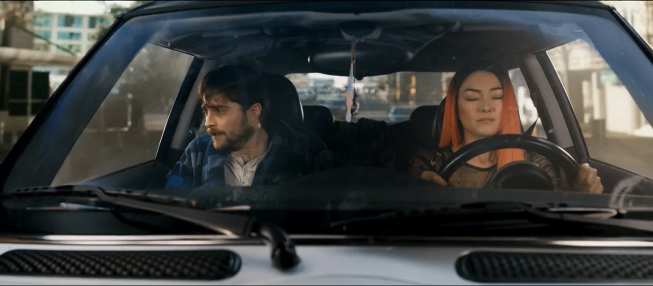Guns Akimbo (2020) movie still: medium — A view from the windshield of a car traveling during the daytime. A man and a woman sit in the front seats…