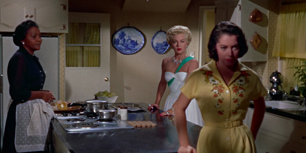 Imitation of Life (1959) movie still: medium — A 1950s home with pastel yellow cabinets and blue decorative plates on the wall. Three women stand…