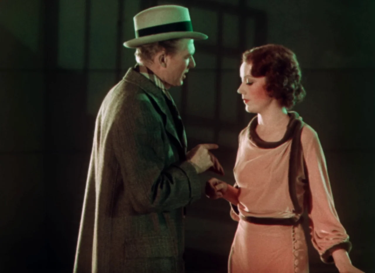 Doctor X (1932) movie still: medium — A dimly lit, abstract studio environment with a dark background and minimal depth. A man in a hat talks to a…