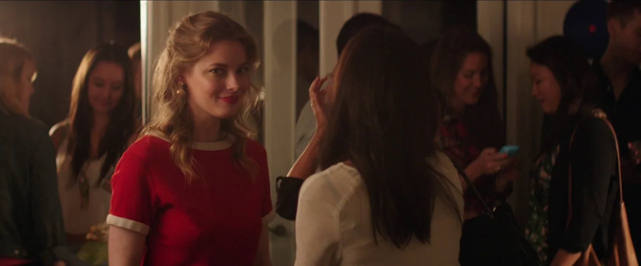 Dean (2016) movie still: medium — A crowded indoor party space with blurred figures in the background. A woman in a red top faces the camera while…