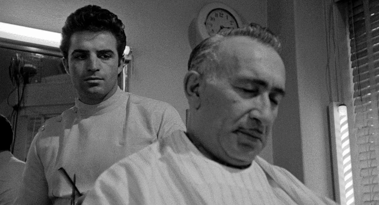 Murder By Contract (1958) movie still: medium — A vintage barbershop interior features a wall clock and a vertical light strip. A young barber in…