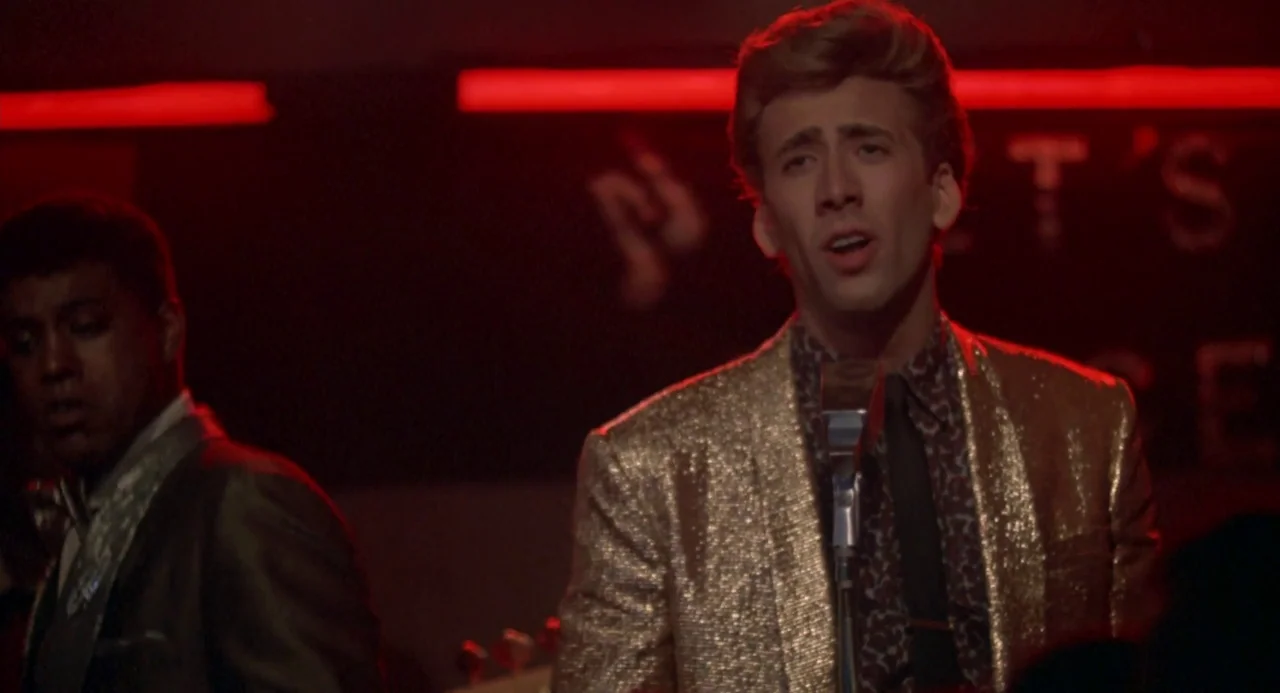 Peggy Sue Got Married (1986) movie still: medium — A dimly lit club interior features a performance stage with vibrant red neon lights. A man in a…