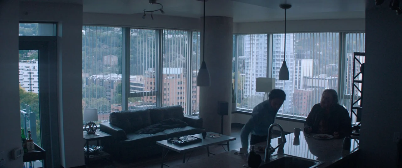 Pig (2021) movie still: wide — A high-rise apartment interior with floor-to-ceiling windows overlooking city buildings. Two men occupy the living…