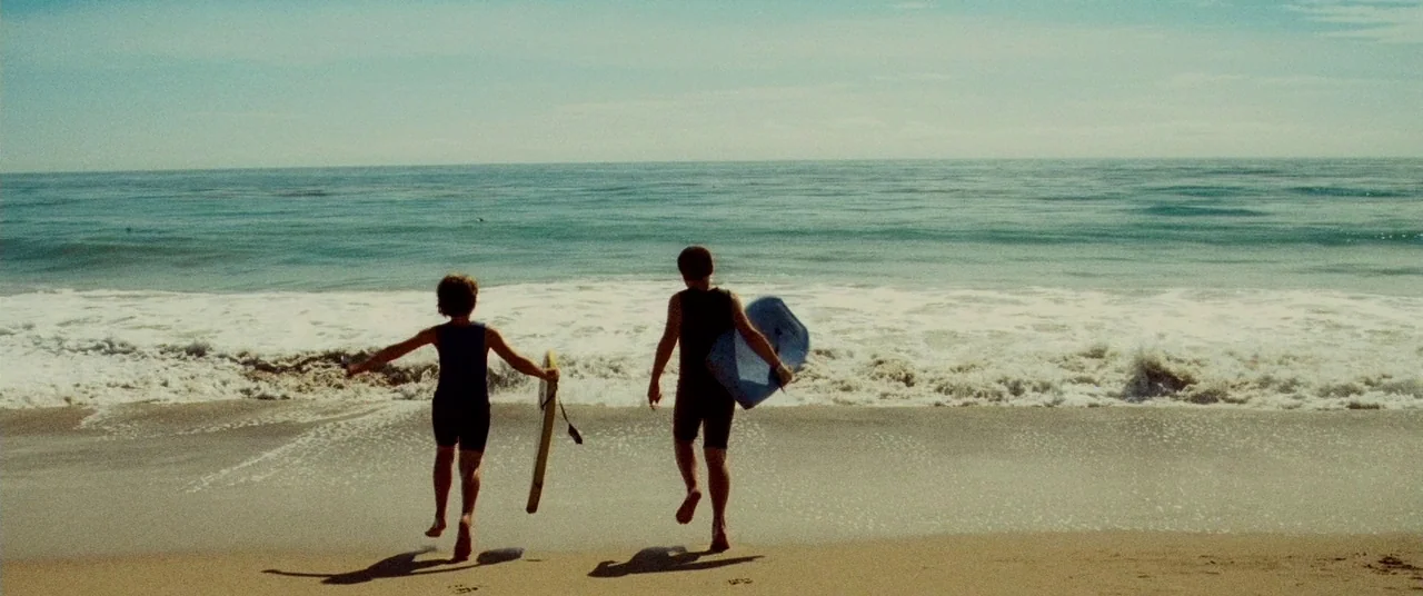 Carriers (2009) movie still: fullbody — A sunny beach with gentle surf and two children running toward the water. They hold boogie boards and their…