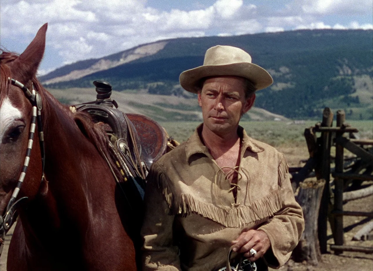 Shane (1953) movie still: medium — A man stands next to a horse in a rural landscape with distant hills. He holds the reins while facing the camera…