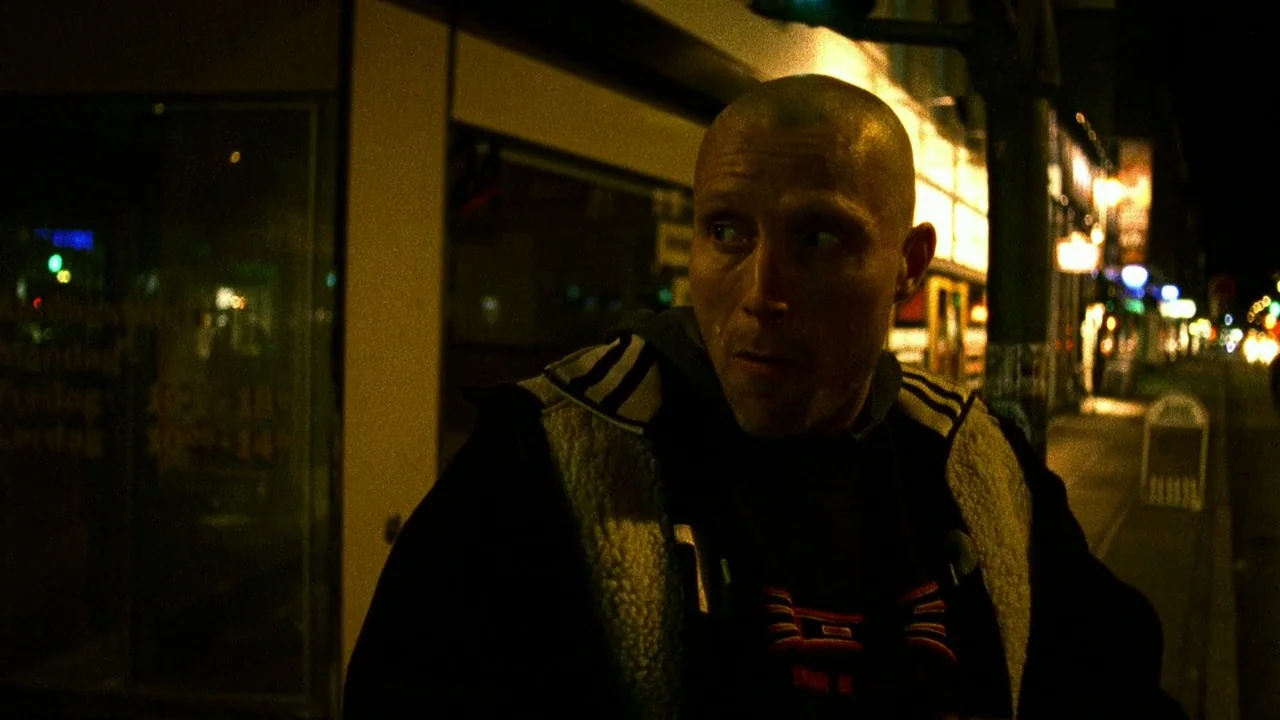 Pusher II (2004) movie still: medium — A nighttime city sidewalk illuminated by harsh, golden-hued artificial lighting from nearby street lamps. A…