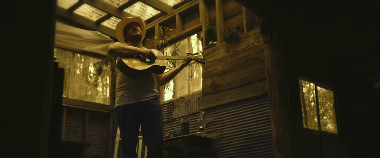 Blaze (2018) movie still: medium — A man plays an acoustic guitar inside a rustic shed with corrugated metal walls. Natural light filters through a…