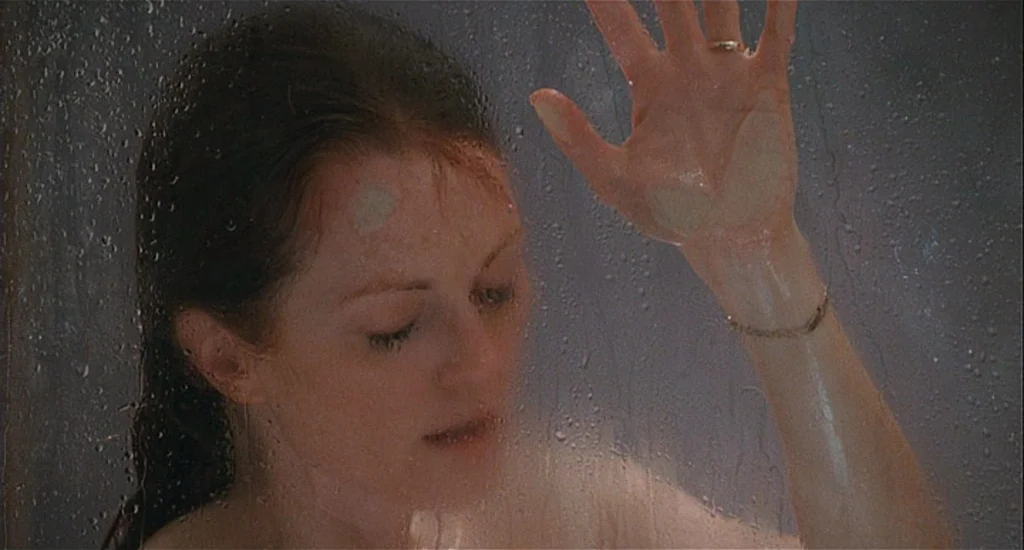 Chloe (2009) movie still: closeup — A woman stands behind a wet, translucent glass wall in an indoor shower stall. She rests one hand against the…