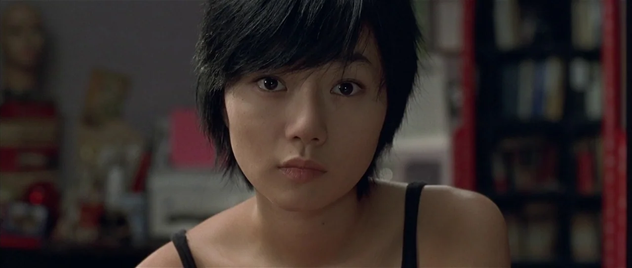 Sympathy for Mr. Vengeance (2002) movie still: closeup — A dimly lit room contains shelves and various personal items in the blurred background. A…