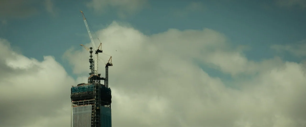 The Report (2019) movie still: establishing — A tall skyscraper under construction stands against a cloudy, overcast daytime sky. Two tower cranes…