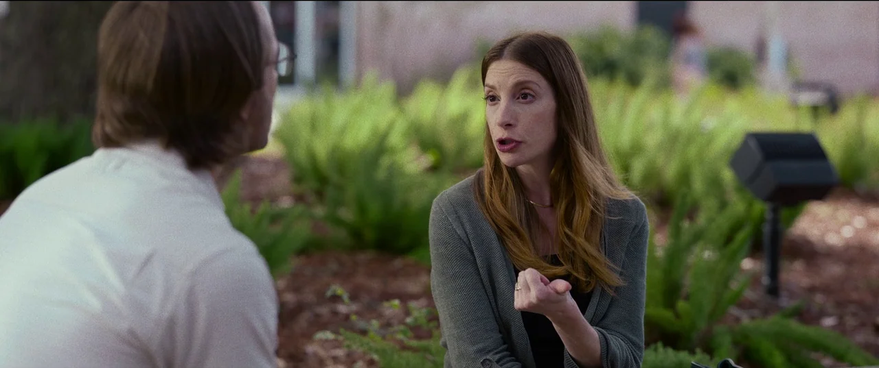 Hit Man (2023) movie still: medium — A man and woman talk outdoors in a landscaped area with ferns and mulch. The woman wears a grey cardigan and…