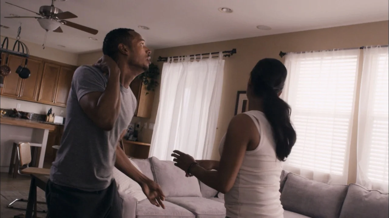 A Haunted House (2013) movie still: medium — A residential living room features light walls, white sheer curtains, and brown wooden cabinetry. A man…