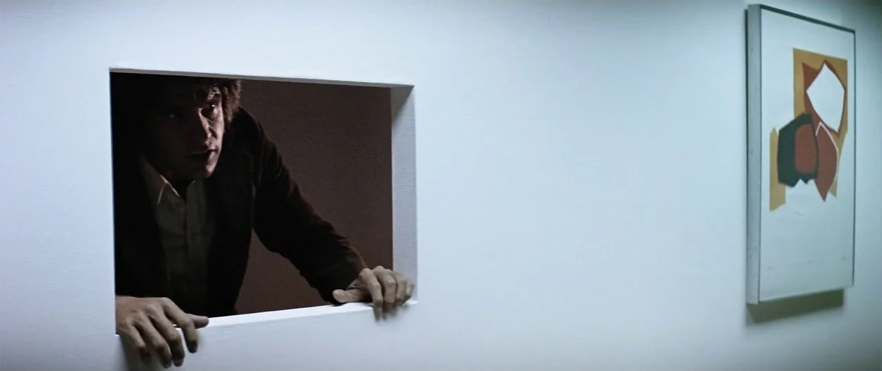 The Parallax View (1974) movie still: medium — A stark white interior wall features a cut-out aperture and an abstract framed print. A man in a…