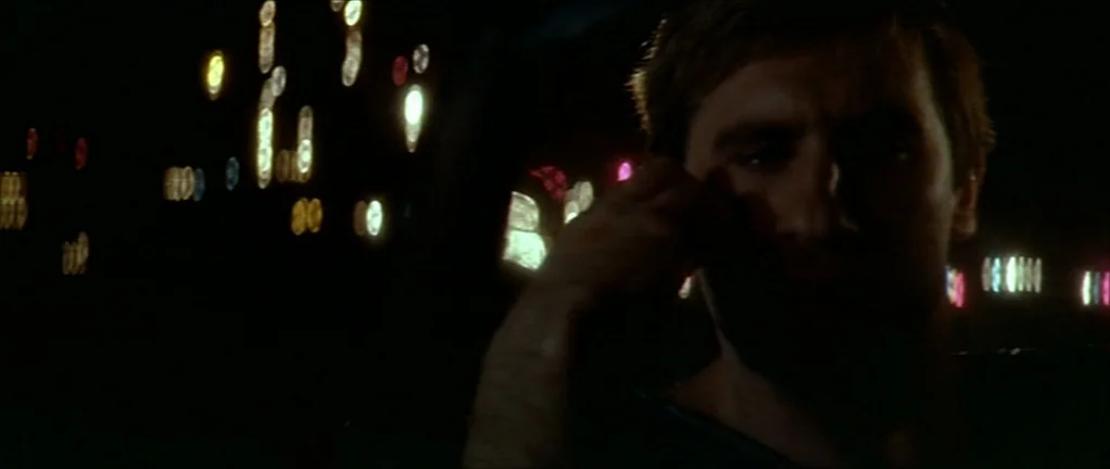 The Moon in the Gutter (1983) movie still: closeup — Inside a dark vehicle at night with blurred city lights in the background. A man sits in the…
